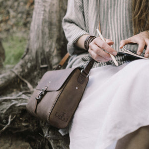 a woman sitting outside wearing a rich brown leather satchel with hand stitched detailing, stainless steel hardware closures and etched logo detailing