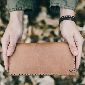 two hands holding a natural tan leather wallet with hand stitching and etched logo details. 