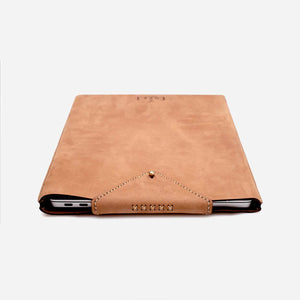a tan brown leather laptop sleeve lying flat on a white surface with a laptop inside. Laptop sleeve has hand stitched and etched logo detailing with a brass hardware closure.