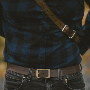 a man standing wearing a rich brown leather belt with an aged brass buckle and etched logo detailing.
