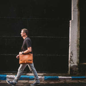 image of a man walking down a street with a large caramel brown leather messenger style bag over one shoulder. 