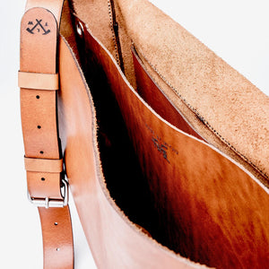 cropped image to reveal inside of a caramel brown messenger style leather handbag with hand stitching, etched logo, and chrome buckle hardware details.
